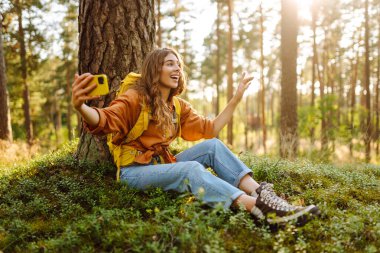 Mutlu genç bir kadın, güneşli bir günde ormanlık alanda bir ağacın yanında otururken selfie çekiyor. Seyahat blogu. Macera, tatil.