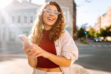 Genç bir kadın, klasik mimariyle çevrili canlı bir şehir caddesinde akıllı telefonunu kullanırken güneşli bir günün tadını çıkarıyor. Teknoloji, blog yazma, iletişim kavramı.