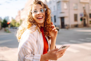 Genç bir kadın, klasik mimariyle çevrili canlı bir şehir caddesinde akıllı telefonunu kullanırken güneşli bir günün tadını çıkarıyor. Teknoloji, blog yazma, iletişim kavramı.