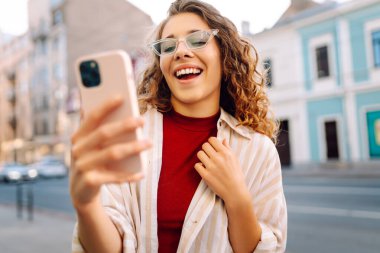 Genç bir kadın, klasik mimariyle çevrili canlı bir şehir caddesinde akıllı telefonunu kullanırken güneşli bir günün tadını çıkarıyor. Teknoloji, blog yazma, iletişim kavramı.