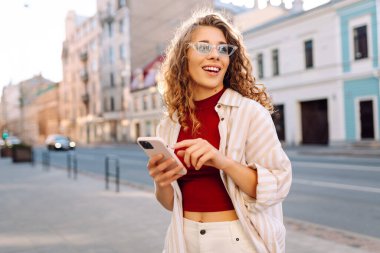 Genç bir kadın, klasik mimariyle çevrili canlı bir şehir caddesinde akıllı telefonunu kullanırken güneşli bir günün tadını çıkarıyor. Teknoloji, blog yazma, iletişim kavramı.