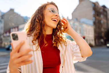 Genç bir kadın, klasik mimariyle çevrili canlı bir şehir caddesinde akıllı telefonunu kullanırken güneşli bir günün tadını çıkarıyor. Teknoloji, blog yazma, iletişim kavramı.