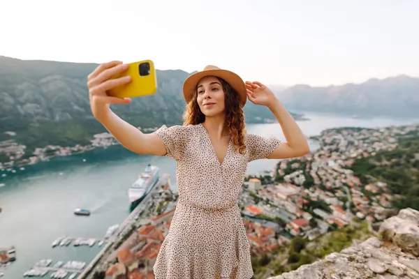 Kıyı kasabasında gün batımında körfezin üstünde manzaranın tadını çıkarırken akıllı telefonuyla selfie çeken genç bir kadın. Seyahat, turizm, teknoloji, blog, iletişim kavramı.