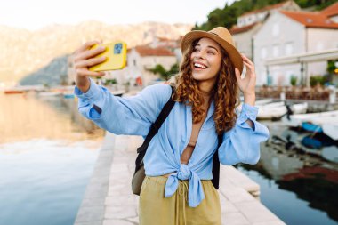 Güneşli bir günde, güzel bir sahil köyünde neşeli bir selfie çeken genç bir kadın. Seyahat, turizm, teknoloji, blog, iletişim kavramı.