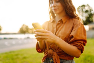 Ellerinizi telefonla kapatın. Suyun kenarında güneşli bir günün tadını çıkaran bir kadın öğleden sonra geç saatlerde parkta akıllı telefonunu kullanıyor. Teknoloji, bağlantı, blog..