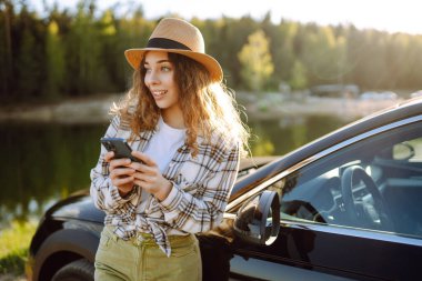 Gölün kenarında güneşli bir günün tadını çıkaran genç bir kadın akşam ışığında park halindeki bir arabanın yanında telefonunu kontrol ediyor. Rav, hafta sonu, doğa, rahatlama ve yaşam tarzı konsepti.
