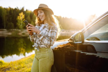Gölün kenarında güneşli bir günün tadını çıkaran genç bir kadın akşam ışığında park halindeki bir arabanın yanında telefonunu kontrol ediyor. Rav, hafta sonu, doğa, rahatlama ve yaşam tarzı konsepti.