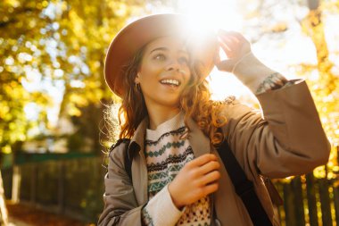 Parkta güneşli bir sonbahar gününün tadını çıkaran genç bir kadın, altın yapraklarla çevriliyken şapkasını takıp gülümsüyor. Seyahat, turizm, hafta sonu