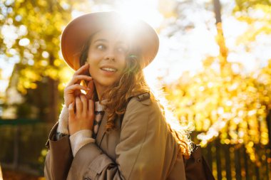 Parkta güneşli bir sonbahar gününün tadını çıkaran genç bir kadın, altın yapraklarla çevriliyken şapkasını takıp gülümsüyor. Seyahat, turizm, hafta sonu