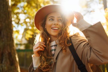 Parkta güneşli bir sonbahar gününün tadını çıkaran genç bir kadın, altın yapraklarla çevriliyken şapkasını takıp gülümsüyor. Seyahat, turizm, hafta sonu
