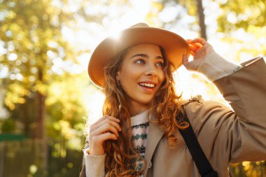 Parkta güneşli bir sonbahar gününün tadını çıkaran genç bir kadın, altın yapraklarla çevriliyken şapkasını takıp gülümsüyor. Seyahat, turizm, hafta sonu