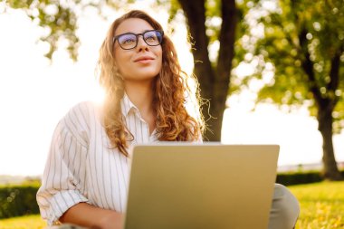 Öğleden sonra güneşli bir parkta dizüstü bilgisayarda çalışan genç bir kadın doğanın ve sıcak havanın tadını çıkarıyor. Çevrimiçi eğitim, Serbest çalışma, teknoloji kavramı.