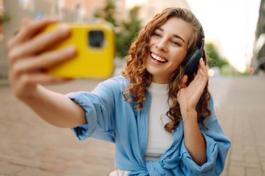 Güneşli bir günde şehir caddesinde kulaklık takıp selfie çeken mutlu genç bir kadın. Kasaba yaşam tarzları. Seyahat, turizm, teknoloji, blog konsepti.