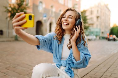 Güneşli bir günde şehir caddesinde kulaklık takıp selfie çeken mutlu genç bir kadın. Kasaba yaşam tarzları. Seyahat, turizm, teknoloji, blog konsepti.