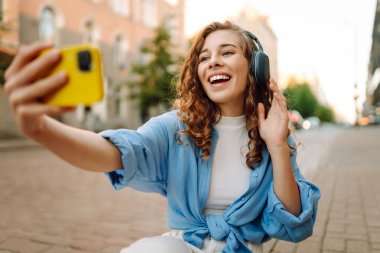 Güneşli bir günde şehir caddesinde kulaklık takıp selfie çeken mutlu genç bir kadın. Kasaba yaşam tarzları. Seyahat, turizm, teknoloji, blog konsepti.
