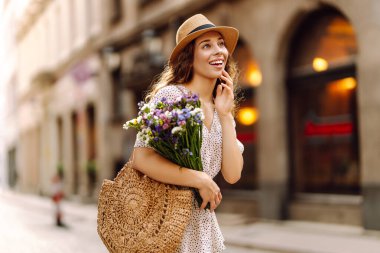Güneşli bir şehirde güneşli bir sokakta gülümserken güneş şapkalı neşeli bir kadın bir buket kır çiçeği tutuyor. Moda tarzı.