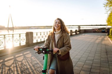 Genç bir kadın, deniz kenarında manzaralı gezintide elektrikli scooterla güneşli bir günün tadını çıkarıyor. Eko-bilinçli taşıma. Seyahat, turizm konsepti.