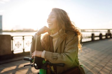 Genç bir kadın, deniz kenarında manzaralı gezintide elektrikli scooterla güneşli bir günün tadını çıkarıyor. Eko-bilinçli taşıma. Seyahat, turizm konsepti.