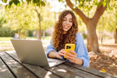 Parktaki ahşap bir masada oturan genç bir kadın elinde akıllı bir telefonla dizüstü bilgisayarına odaklanmış. Eğitim ve modern teknolojiler.