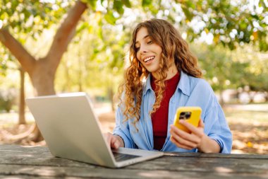 Parktaki ahşap bir masada oturan genç bir kadın elinde akıllı bir telefonla dizüstü bilgisayarına odaklanmış. Eğitim ve modern teknolojiler.