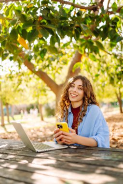 Parktaki ahşap bir masada oturan genç bir kadın elinde akıllı bir telefonla dizüstü bilgisayarına odaklanmış. Eğitim ve modern teknolojiler.