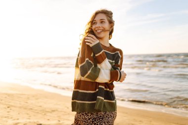 Kumsalda günbatımının tadını çıkaran genç bir kadın rahat çizgili bir süveter giyerken, dalgalar yavaşça kıyıya vururken gülümsüyor. Seyahat, hafta sonu, rahatlama ve yaşam tarzı konsepti.