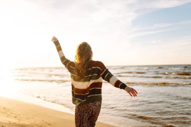 Kumsalda günbatımının tadını çıkaran genç bir kadın rahat çizgili bir süveter giyerken, dalgalar yavaşça kıyıya vururken gülümsüyor. Seyahat, hafta sonu, rahatlama ve yaşam tarzı konsepti.