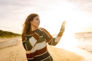 Kumsalda günbatımının tadını çıkaran genç bir kadın rahat çizgili bir süveter giyerken, dalgalar yavaşça kıyıya vururken gülümsüyor. Seyahat, hafta sonu, rahatlama ve yaşam tarzı konsepti.