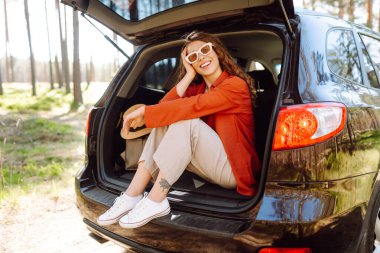 Bir gezgin genç kadın yemyeşil yemyeşil bir ormanda güneşli bir günde arabasının bagajında otururken gülümsüyor. Yaz tatili seyahatleri..
