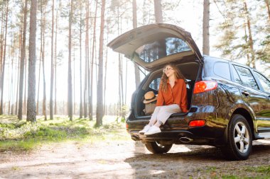 Bir gezgin genç kadın yemyeşil yemyeşil bir ormanda güneşli bir günde arabasının bagajında otururken gülümsüyor. Yaz tatili seyahatleri..