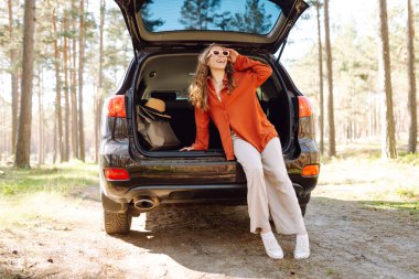 Bir gezgin genç kadın yemyeşil yemyeşil bir ormanda güneşli bir günde arabasının bagajında otururken gülümsüyor. Yaz tatili seyahatleri..