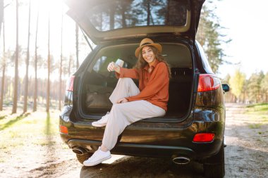 Bir gezgin genç kadın yemyeşil yemyeşil bir ormanda güneşli bir günde arabasının bagajında otururken gülümsüyor. Yaz tatili seyahatleri..