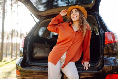 Bir gezgin genç kadın yemyeşil yemyeşil bir ormanda güneşli bir günde arabasının bagajında otururken gülümsüyor. Yaz tatili seyahatleri..