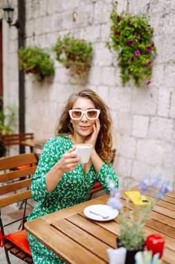 Güneşli bir öğleden sonra yeşil çiçekli bir elbise giymiş güzel bir kasabada, açık bir kafede kahve içen bir kadın. İnsanlar, moda, yaşam tarzı, seyahat ve tatiller