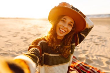 Gün batımında, şapkası ve çizgili süveteri ile kumlu bir plajda neşeli bir selfie çeken genç bir kadın. Teknoloji ve yaşam tarzı seyahat ve seyahat kavramı