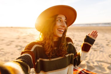 Gün batımında, şapkası ve çizgili süveteri ile kumlu bir plajda neşeli bir selfie çeken genç bir kadın. Teknoloji ve yaşam tarzı seyahat ve seyahat kavramı