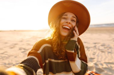 Gün batımında, şapkası ve çizgili süveteri ile kumlu bir plajda neşeli bir selfie çeken genç bir kadın. Teknoloji ve yaşam tarzı seyahat ve seyahat kavramı