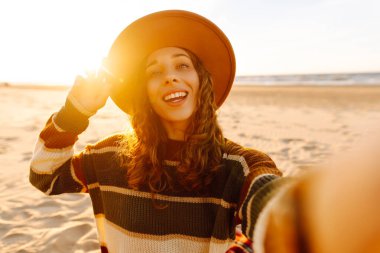 Gün batımında, şapkası ve çizgili süveteri ile kumlu bir plajda neşeli bir selfie çeken genç bir kadın. Teknoloji ve yaşam tarzı seyahat ve seyahat kavramı