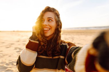 Gün batımında, şapkası ve çizgili süveteri ile kumlu bir plajda neşeli bir selfie çeken genç bir kadın. Teknoloji ve yaşam tarzı seyahat ve seyahat kavramı