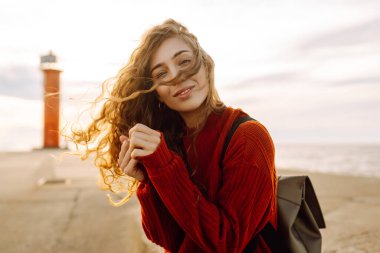 Genç bir kadın, öğleden sonra saatlerinde canlı kırmızı bir deniz fenerinin yanında kumsalda rüzgarlı bir günün tadını çıkarıyor. Seyahat, hafta sonu, rahatlama ve yaşam tarzı konsepti.