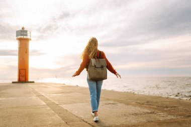 Genç bir kadın, öğleden sonra saatlerinde canlı kırmızı bir deniz fenerinin yanında kumsalda rüzgarlı bir günün tadını çıkarıyor. Seyahat, hafta sonu, rahatlama ve yaşam tarzı konsepti.