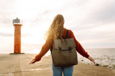 Genç bir kadın, öğleden sonra saatlerinde canlı kırmızı bir deniz fenerinin yanında kumsalda rüzgarlı bir günün tadını çıkarıyor. Seyahat, hafta sonu, rahatlama ve yaşam tarzı konsepti.