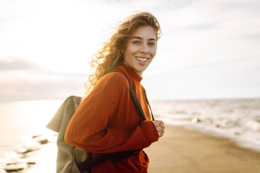 Genç bir kadın, öğleden sonra saatlerinde canlı kırmızı bir deniz fenerinin yanında kumsalda rüzgarlı bir günün tadını çıkarıyor. Seyahat, hafta sonu, rahatlama ve yaşam tarzı konsepti.