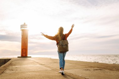 Genç bir kadın, öğleden sonra saatlerinde canlı kırmızı bir deniz fenerinin yanında kumsalda rüzgarlı bir günün tadını çıkarıyor. Seyahat, hafta sonu, rahatlama ve yaşam tarzı konsepti.