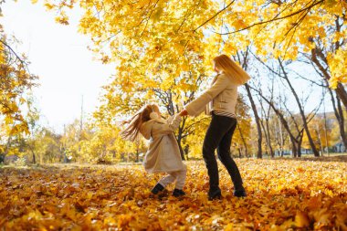 Parkta neşeli bir sonbahar günü. Bir anne ve kızı, parlak sarı ağaçlarla çevrili düşmüş yapraklarda oynuyorlar. Dinlen, çocukluk, yürüyüş, aile.