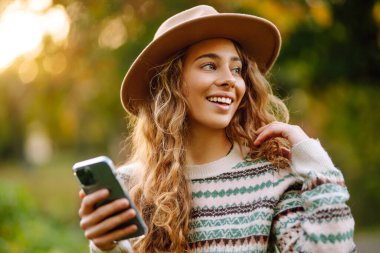 Süveter ve şapka giyen neşeli genç bir kadın renkli yapraklarla dolu bir parkta güneşli bir sonbahar gününün tadını çıkarıyor. Sonbahar kadını. 