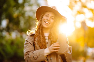 Süveter ve şapka giyen neşeli genç bir kadın renkli yapraklarla dolu bir parkta güneşli bir sonbahar gününün tadını çıkarıyor. Sonbahar kadını. 