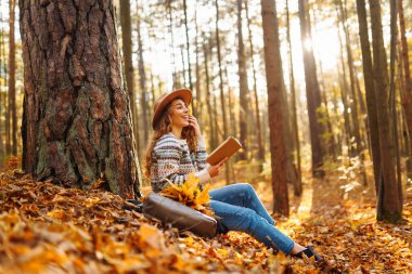 Sonbahar yapraklarının üzerinde otururken kitap okuyan bir kadın altın gün ışığında sakin bir ormanda bisikletinin yanında oturuyor.
