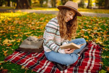 Kıvırcık saçlı bir kadın piknik paspasına oturmuş, canlı sonbahar yaprakları arasında kitap okumaya dalmış. Parkın güzelliği onun huzurlu anını güzelleştirir..