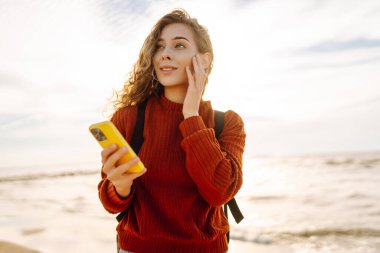 Gün batımında kumsalda gülümseyen ve akıllı telefonunu kullanan genç bir kadın. Altın güneş ışığı saçlarını ve çevresini aydınlatıyor. Bloglama, iletişim konsepti.